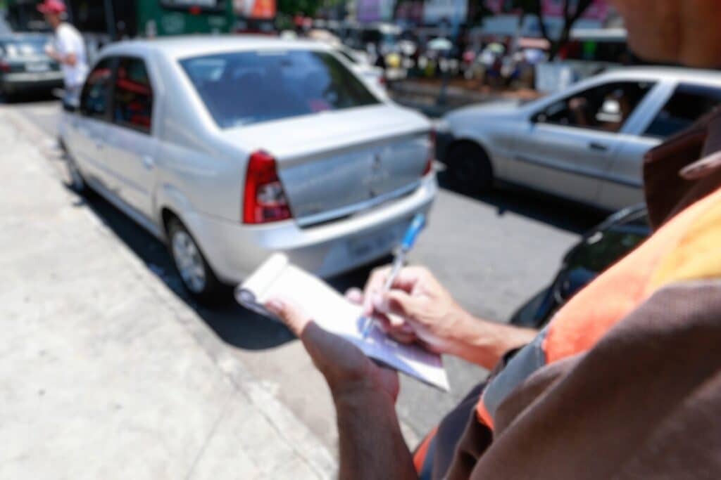 Agente escreve multa em carro estacionado na rua.